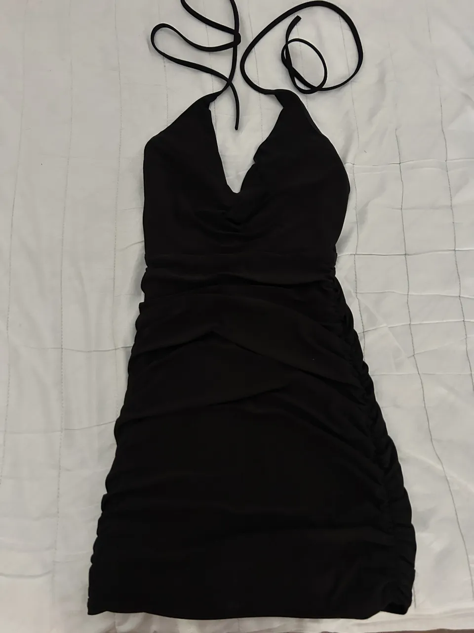 Vestido negro súper canchero, ideal para cualquier salida. Tiene un escote halter con tiras finas que se atan en la nuca y un detalle fruncido en la parte delantera. La falda es ajustada y tiene un efecto fruncido en los laterales que estiliza la figura. ¡Un básico que no puede faltar en tu guardarropa!