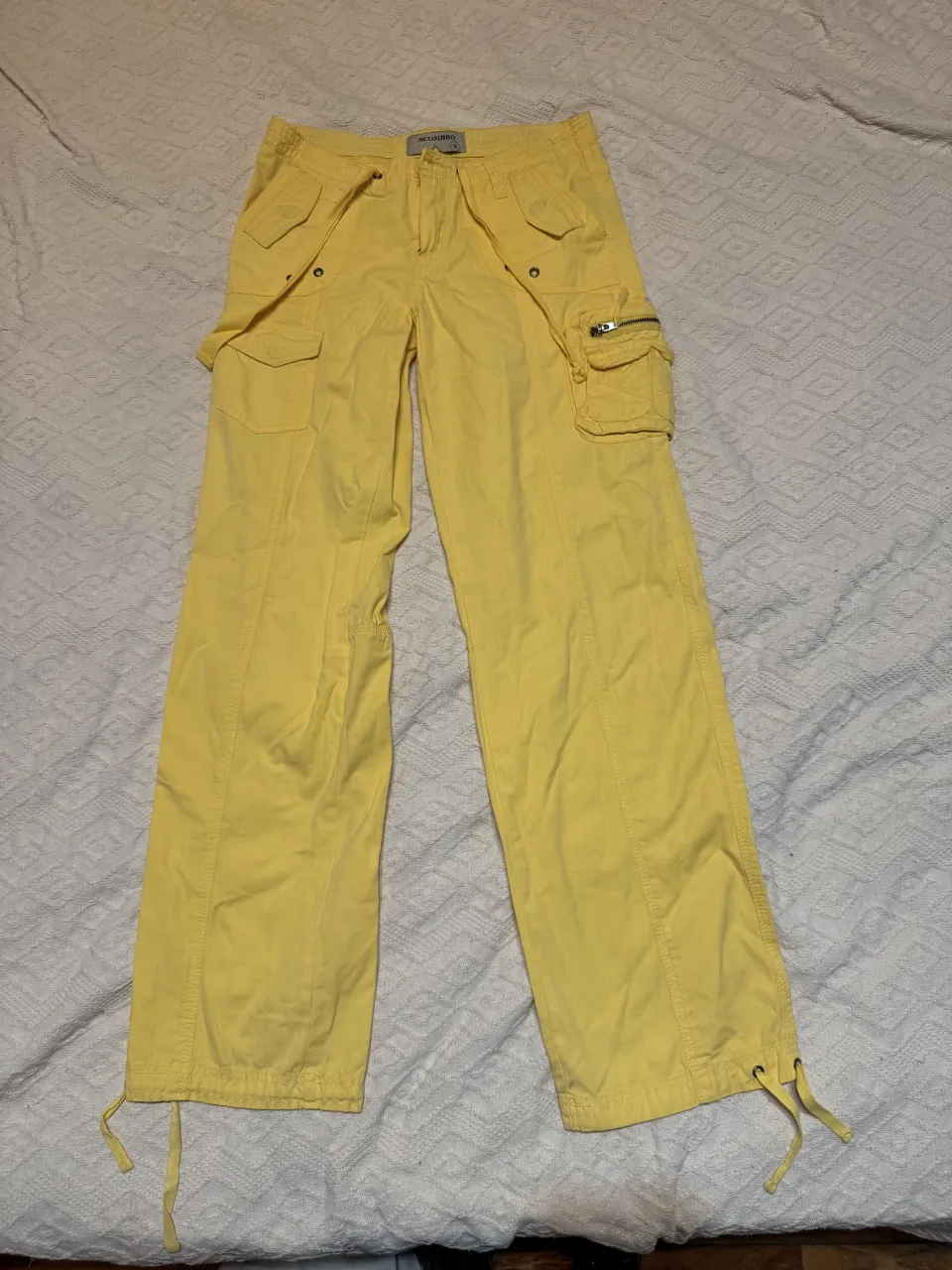 Pantalón cargo amarillo con bolsillos frontales y laterales, uno de ellos con cierre. Posee cordones en los tobillos para ajustar.