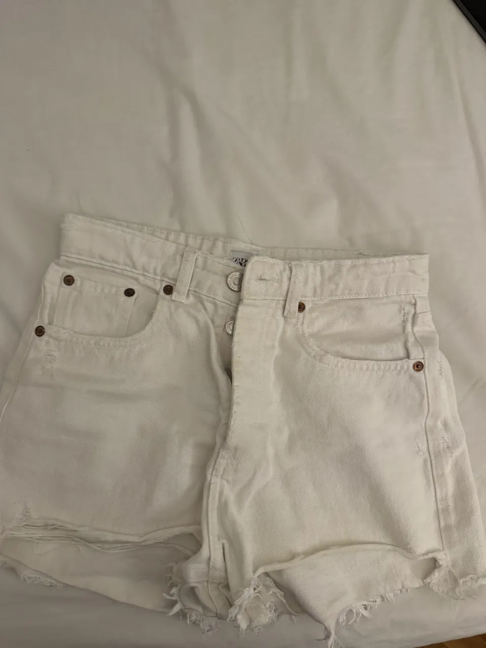 Shorts de jean blancos con roturas y detalles deshilachados en los bordes inferiores. Tienen bolsillos delanteros y traseros.