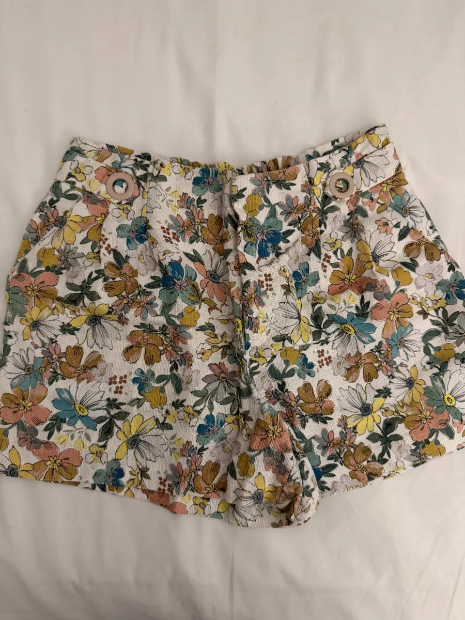 Shorts blancos con estampado floral multicolor. Tienen dos botones en la parte delantera y cintura elastizada.