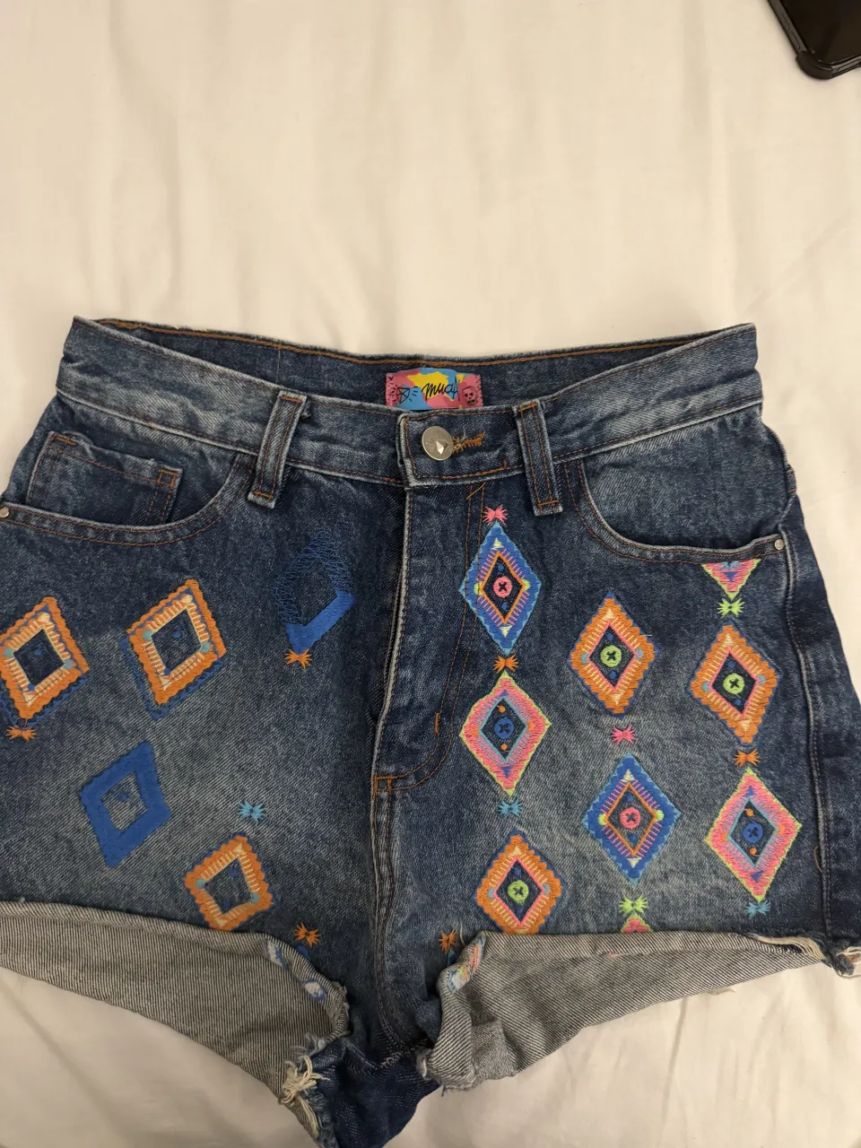 Shorts de jean tiro alto con bordados de rombos y detalles de estrellas en la parte delantera. Posee roturas y terminación deshilachada en el borde inferior.