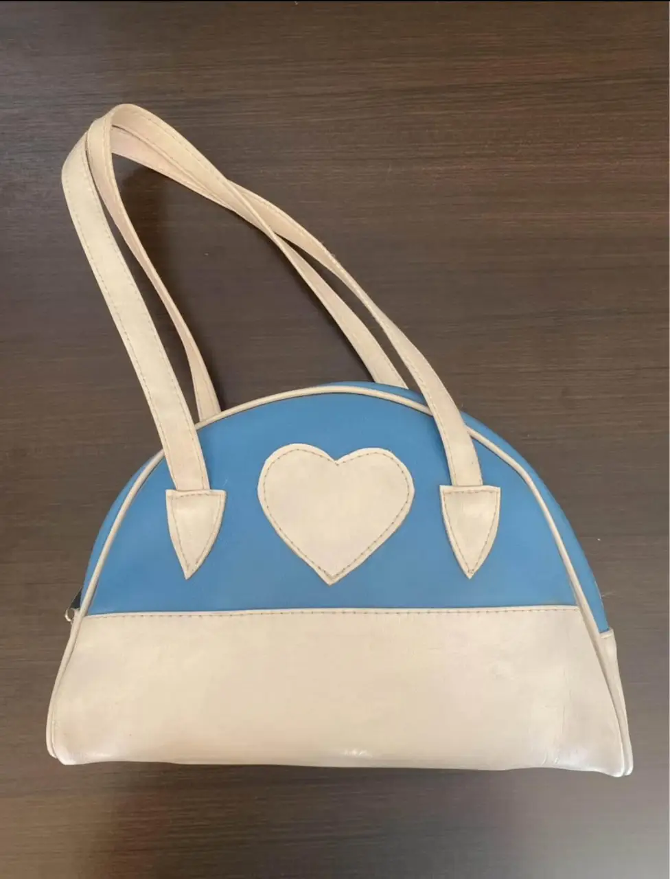 Cartera de mano con forma de media luna, con detalle de corazón en el frente. Combina el azul con el blanco, ideal para un look casual y con onda.