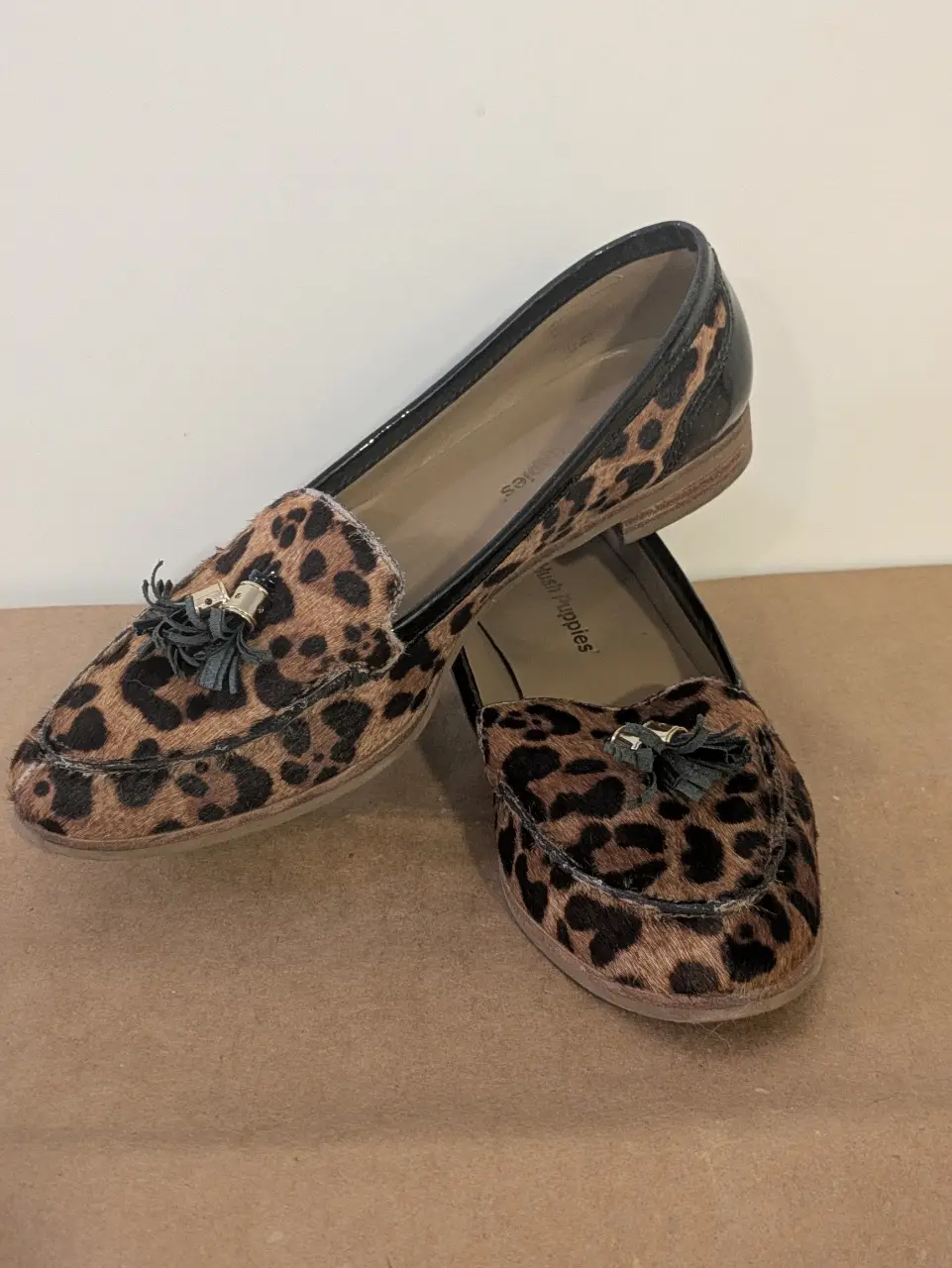 Mocasines de mujer con estampado de leopardo y detalles en charol negro. Tienen borlas decorativas y son de la marca Hush Puppies. Ideales para un look casual y elegante.