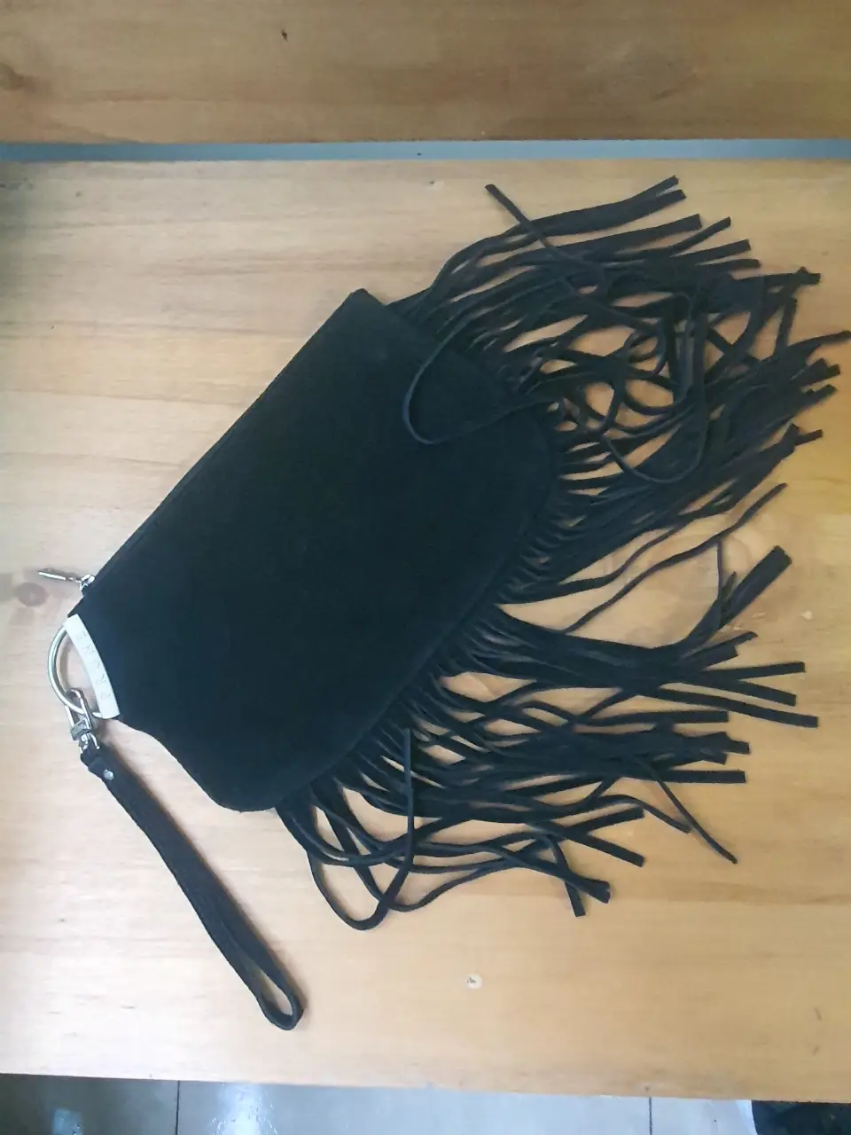 Cartera de mano negra con flecos, ideal para un look bohemio o de fiesta. Confeccionada en gamuza, cuenta con un detalle de argolla y correa para llevarla cómodamente. Interior con forro y detalles de la marca Prune.