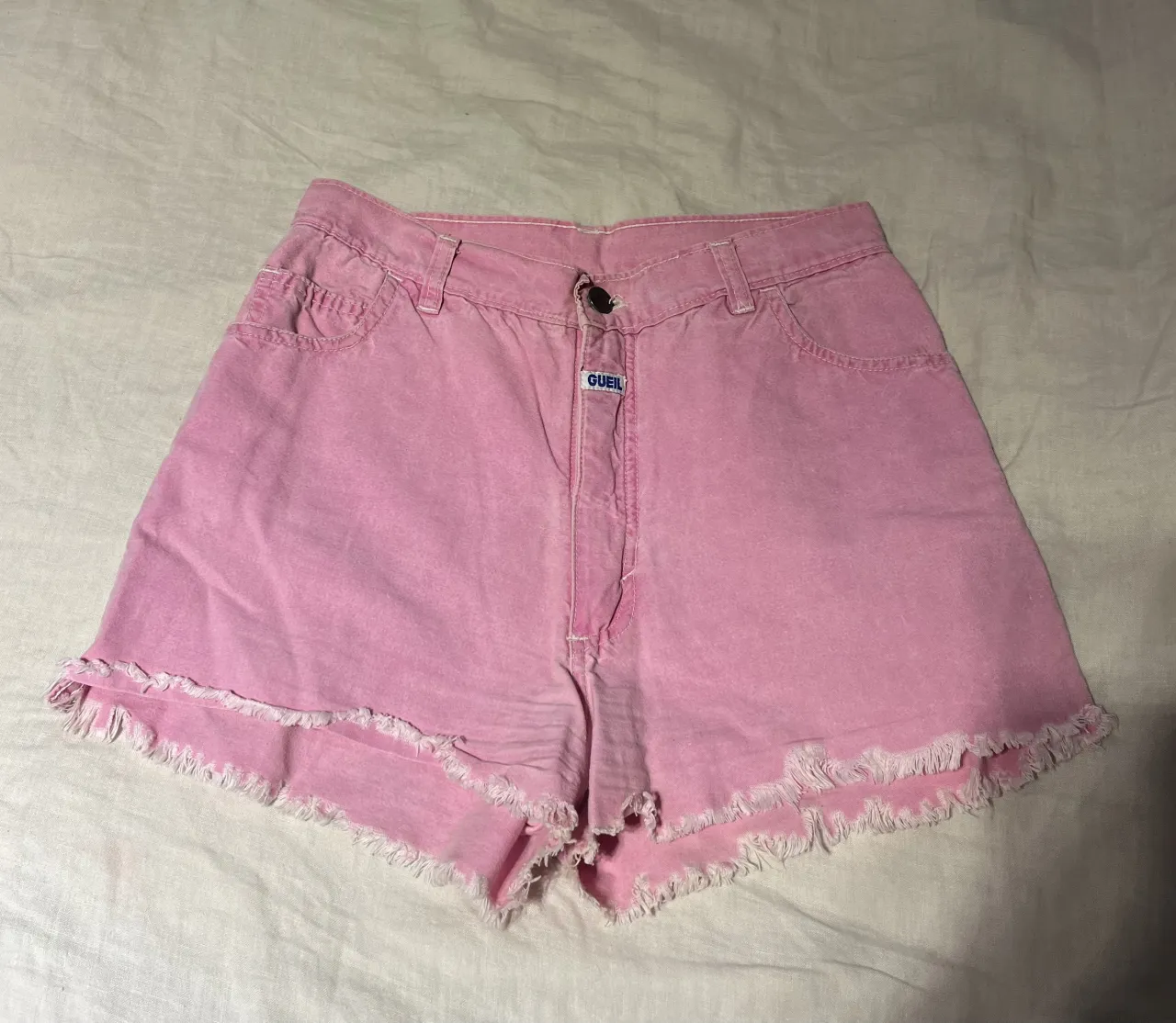 Shorts de jean color rosa con roturas y flecos en el ruedo. Cierre de botón y detalle de etiqueta con logo en la parte delantera.