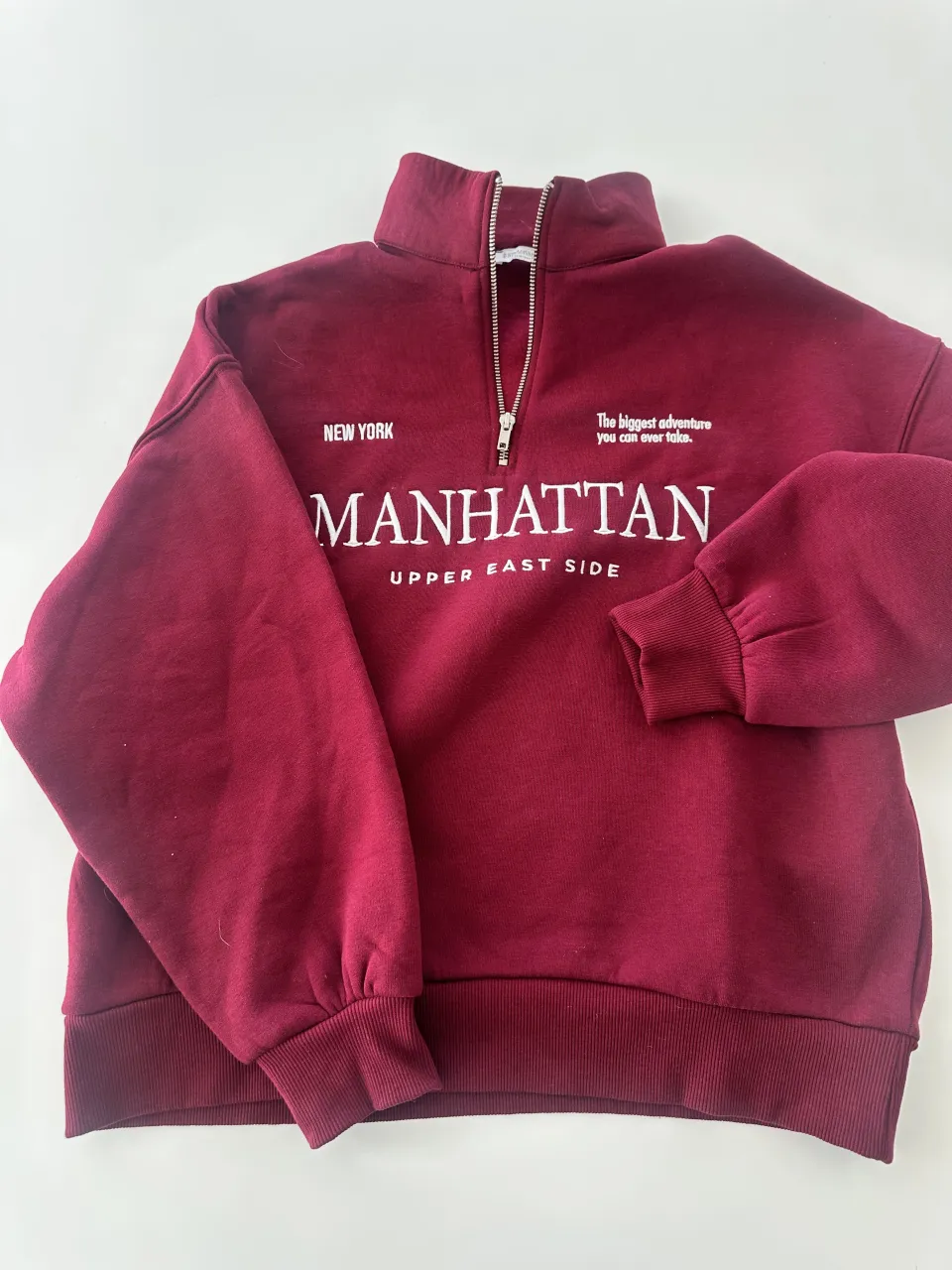 Buzo de color bordo con cierre en el cuello y estampa "MANHATTAN UPPER EAST SIDE" en blanco.