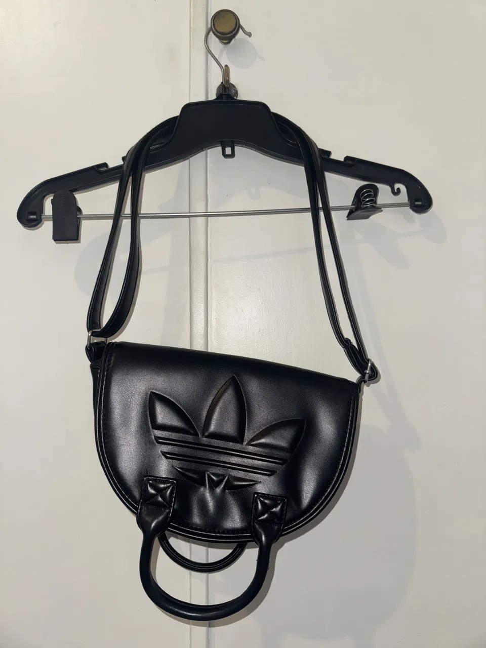 Cartera negra de cuero sintético con logo de Adidas en relieve. Tiene dos asas cortas y una correa larga desmontable. Se cierra con broche.