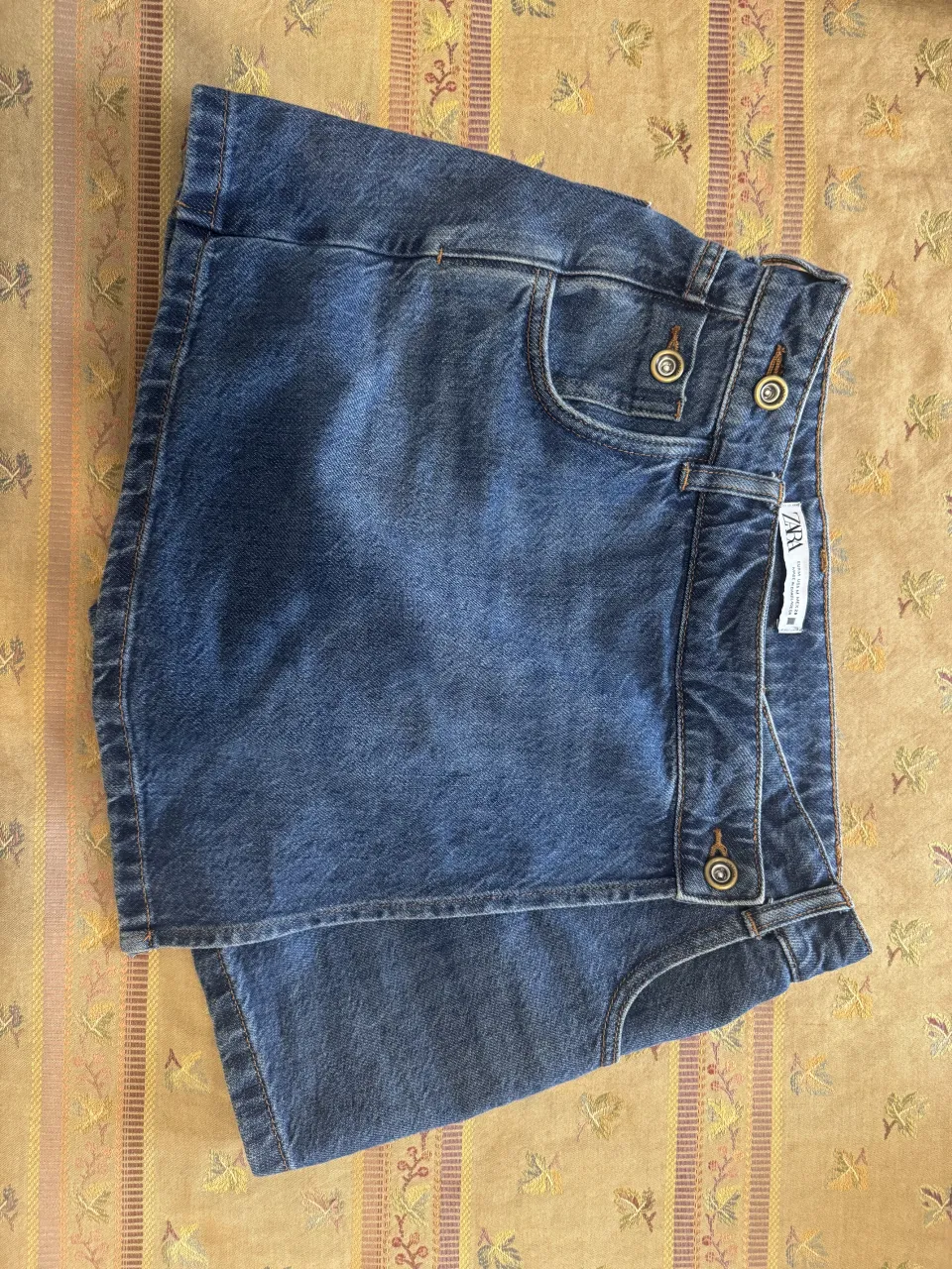 Shorts de jean azul con detalles de costura en marrón. Tienen dos bolsillos delanteros y dos traseros. Cierre con botones.