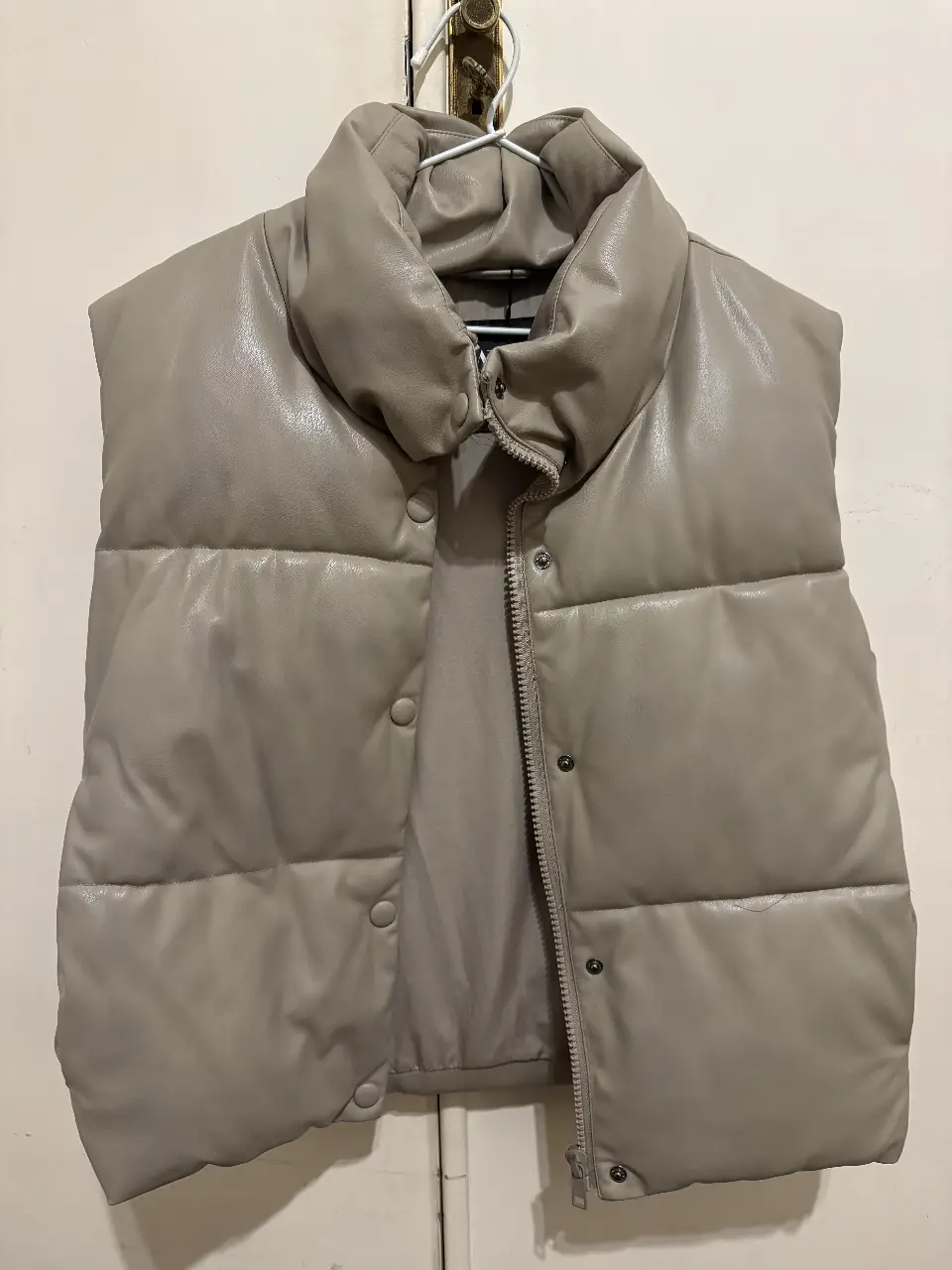 Chaleco puffer de cuero sintético en color gris. Cuello alto y cierre con cremallera y botones. Ideal para un look moderno y abrigado.