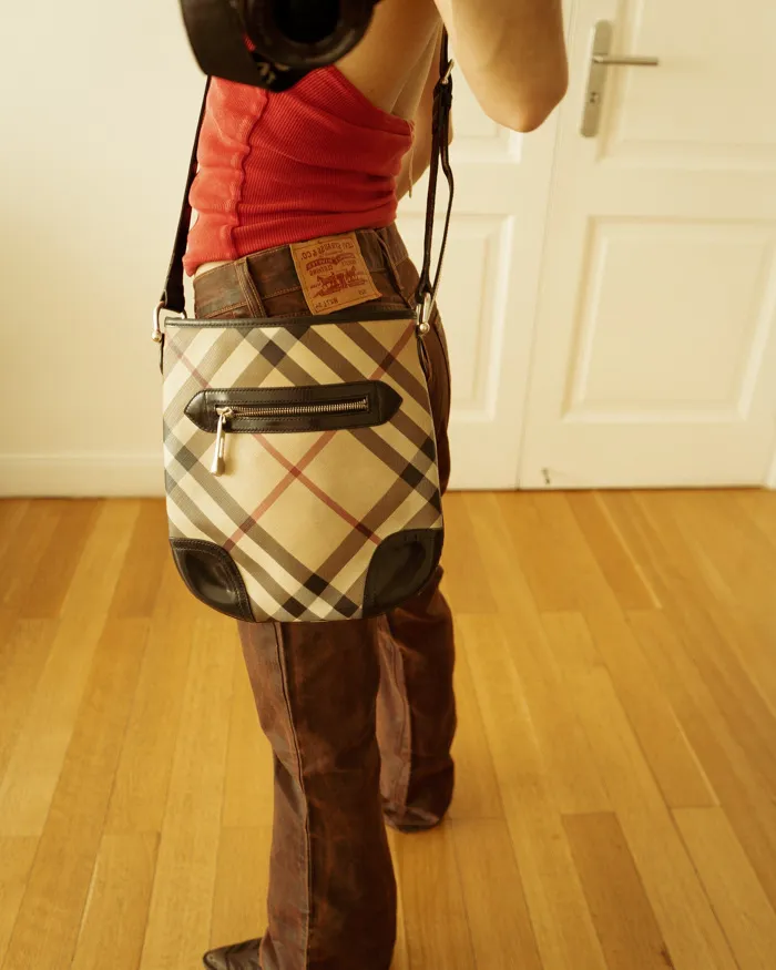 Bandolera Burberry (original) con estampado a cuadros en tonos beige, marrón y rojo. Detalles en cuero negro y cierre con cremallera. 