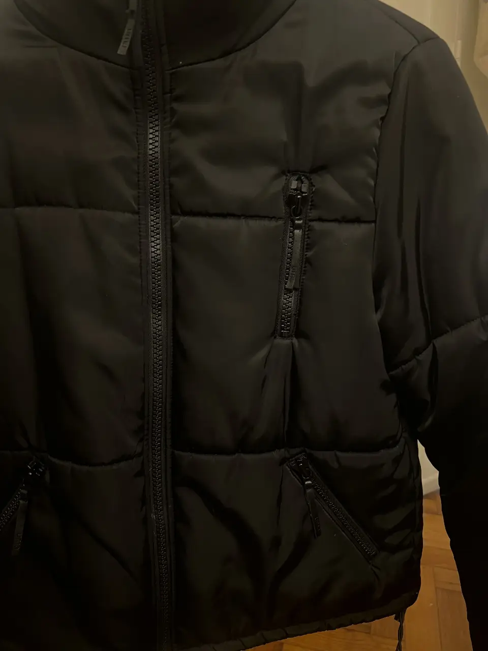 Campera puffer negra, con cierre frontal y bolsillos con cierre. Cuello alto y capucha guardada para lluvia.
