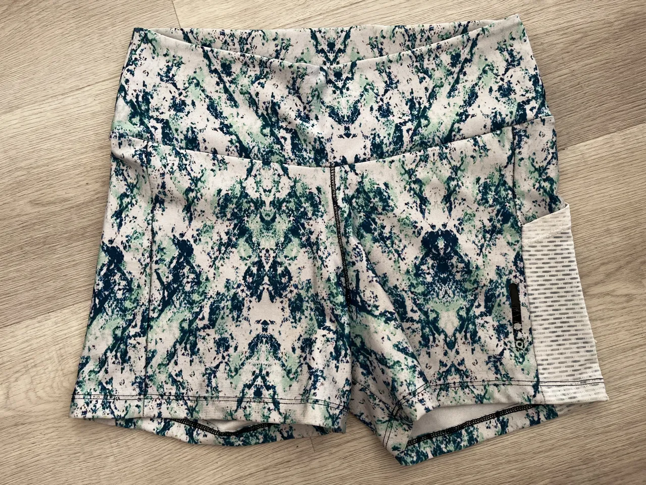 Shorts deportivos con estampado abstracto en tonos verde y azul sobre fondo blanco. Posee un bolsillo lateral de mesh blanco.