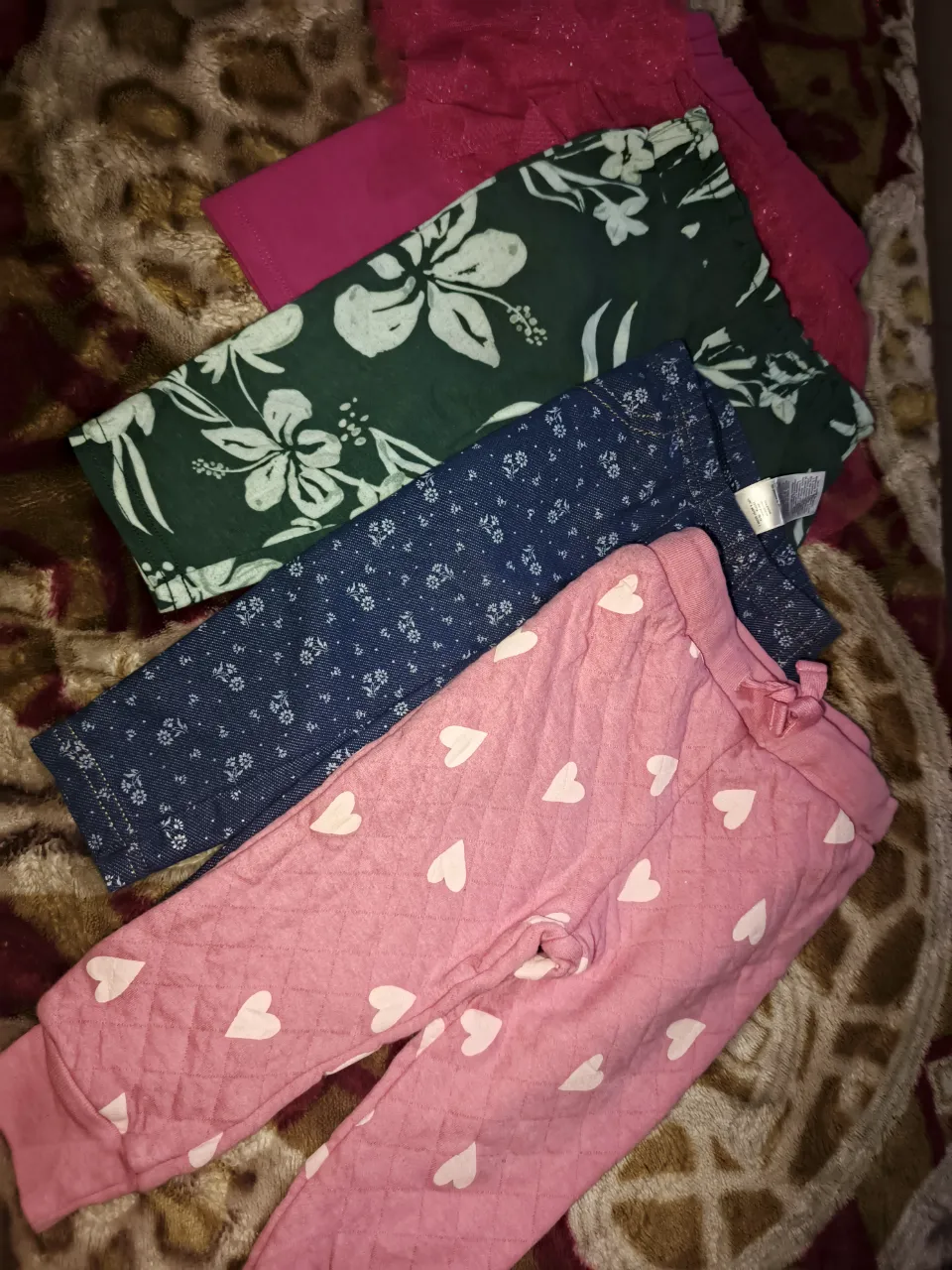 Conjunto de ropa para bebé que incluye varios pantalones y un body. Un pantalón es rosa acolchado con corazones blancos. Otro pantalón es azul con estampado floral pequeño. Hay un pantalón verde con estampado de flores grandes. También se observa un body rojo con estampado floral y un body blanco con texto 'MOMMY'S MINI' junto a un short verde con estampado de mariposas. Finalmente, un mameluco a cuadros multicolores con pies.