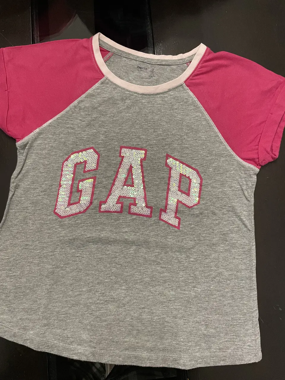 Remera de manga corta para niña, con mangas y cuello en color rosa. El frente presenta el logo "GAP" en lentejuelas plateadas. Ideal para un look casual y cómodo.