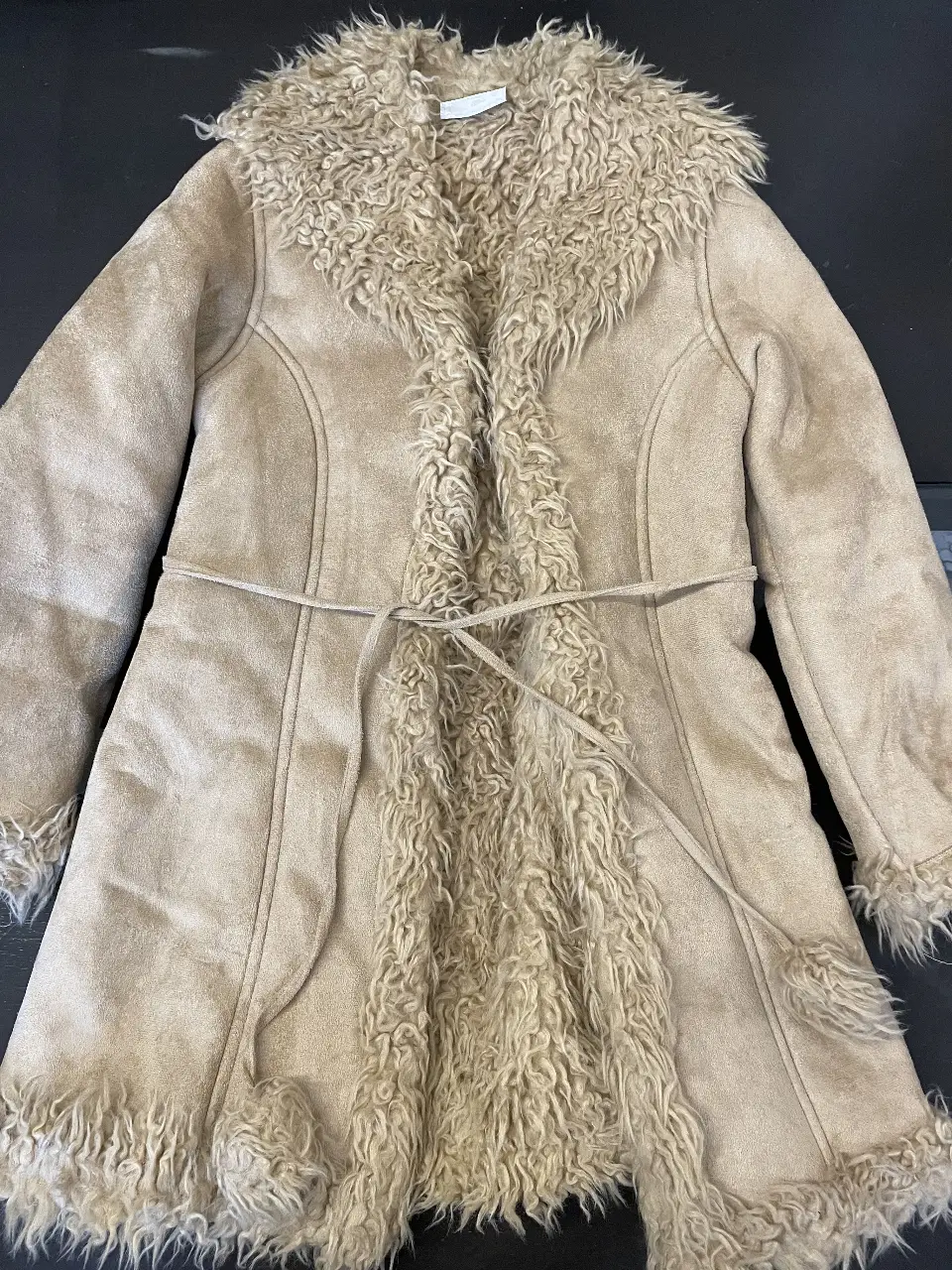 Tapado largo de gamuza sintética color crudo con detalles de piel sintética en cuello, solapa, borde y puños. Incluye cinturón para ajustar la cintura. Ideal para un look boho chic.