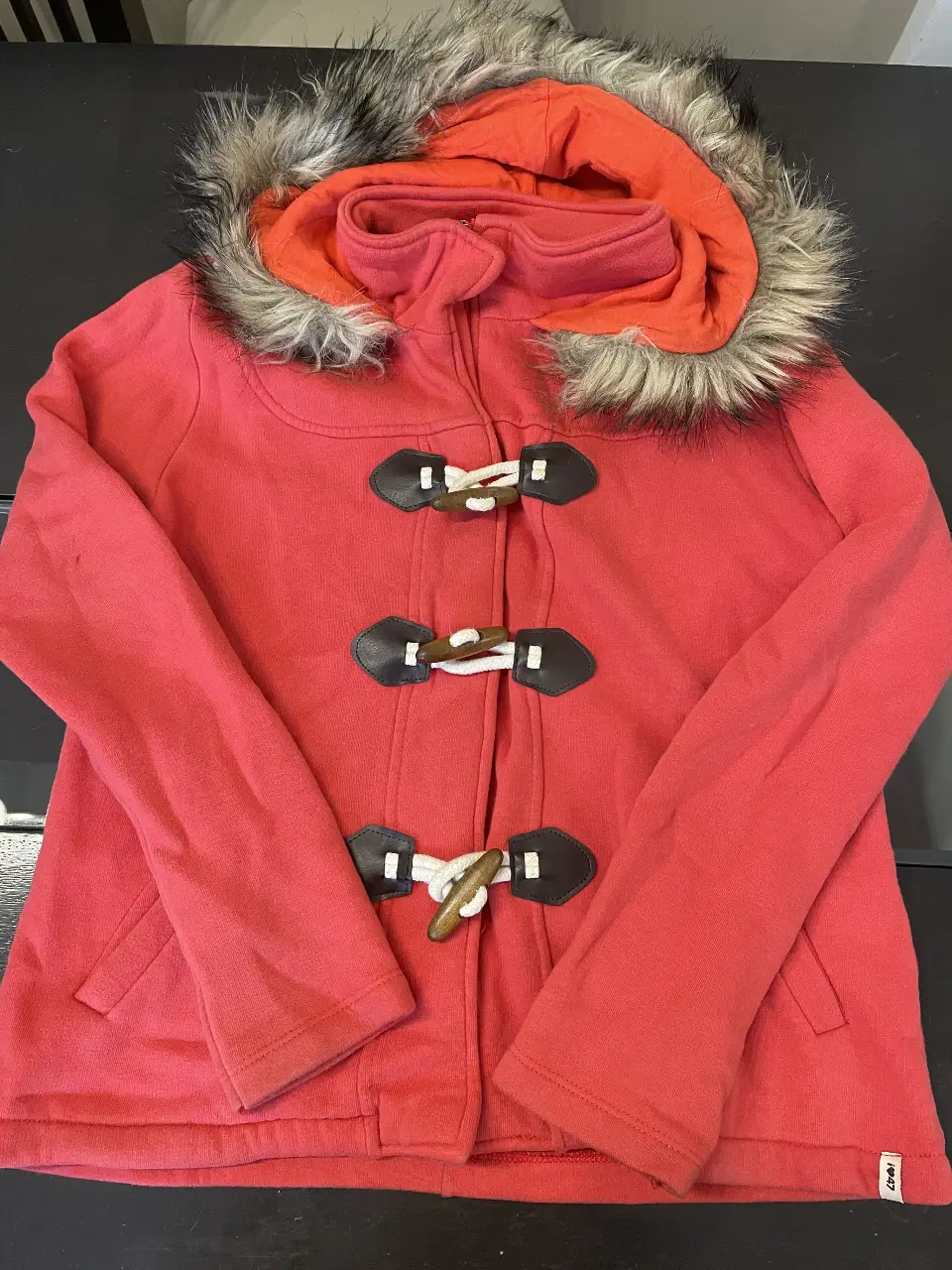 Campera roja con capucha y borde de piel sintética. Cierre frontal y detalles de madera en los broches. Ideal para el invierno.