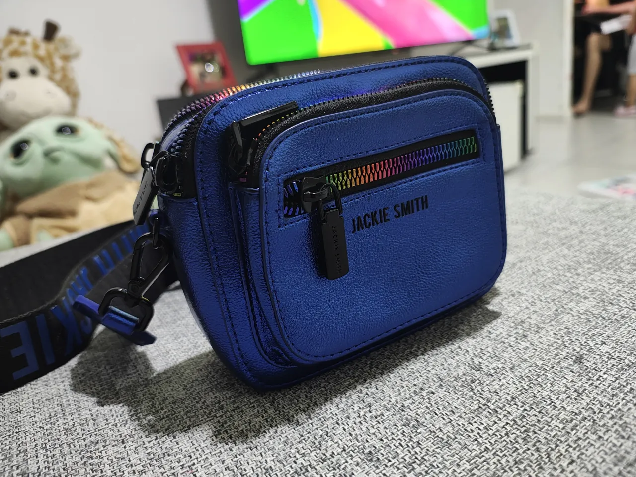 Bolso bandolera azul metálico de la marca Jackie Smith. Tiene múltiples compartimentos con cierres de arcoíris y correa ajustable con el logo de la marca. Ideal para el día a día o para darle un toque moderno a tu outfit.