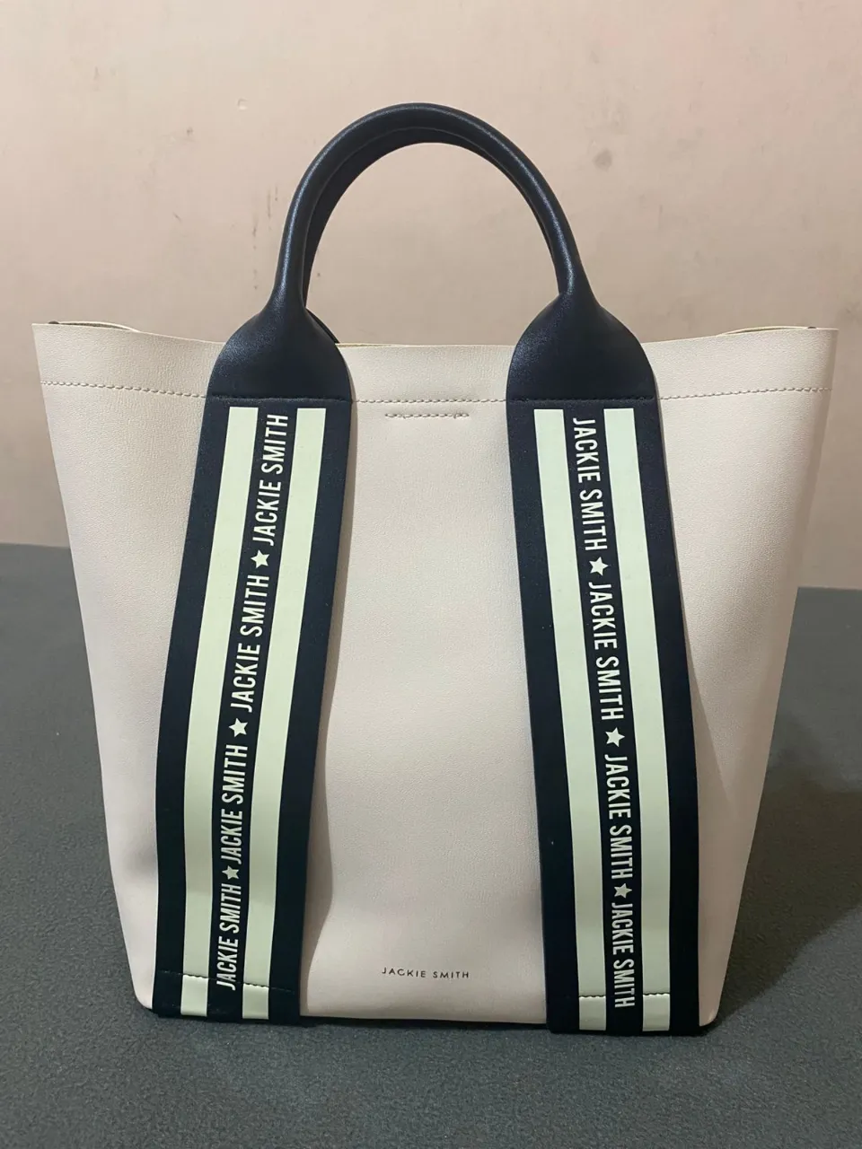 Bolso tote de cuero color crudo, con tiras negras estampadas con el logo de la marca "Jackie Smith" en blanco y detalles en amarillo. Incluye correa desmontable. ¡Un clásico para tu día a día!