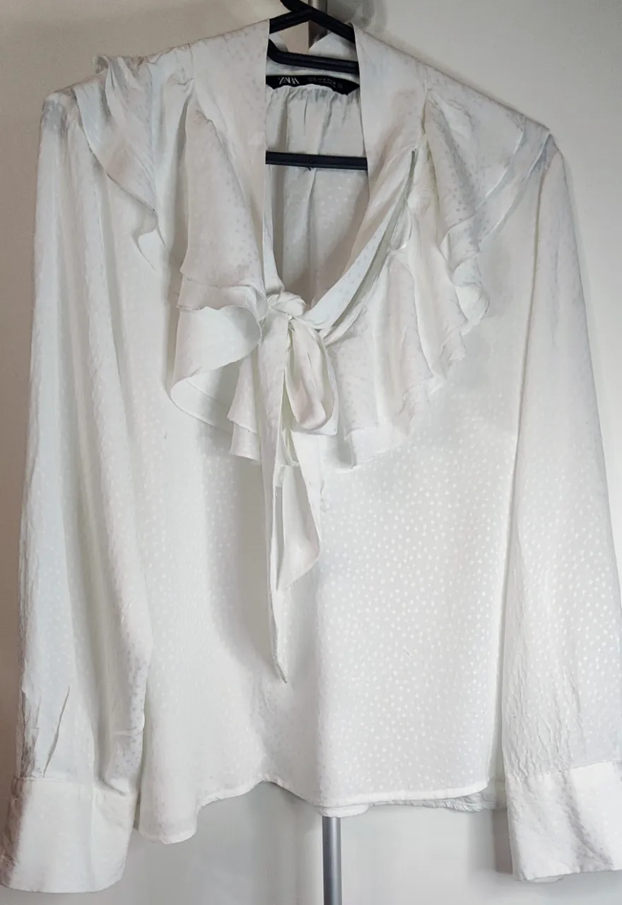 Blusa blanca romántica de la marca Zara, caracterizada por su tejido con textura  plumeti o jacquard con pequeños puntos y un diseño detallado con volantes y lazada, un cuello tipo Lavallière con una lazada frontal que permite ajustarla al gusto, que caen desde los hombros hacia el pecho, aportando un estilo bohemio y elegante. b Posee botones con  en los puños. de sisa a sisa 50 cm.