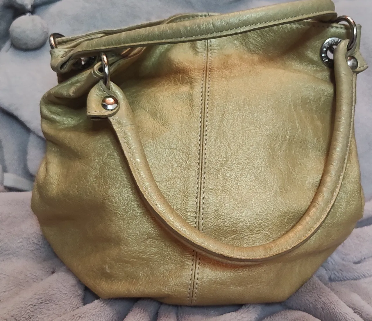 cartera dorada de cuero presenta un diseño de estilo hobo o "bolsito" con un acabado metalizado y detalles de herrajes circulares, de cuero dorado con manijas cortas y detalles metálicos.