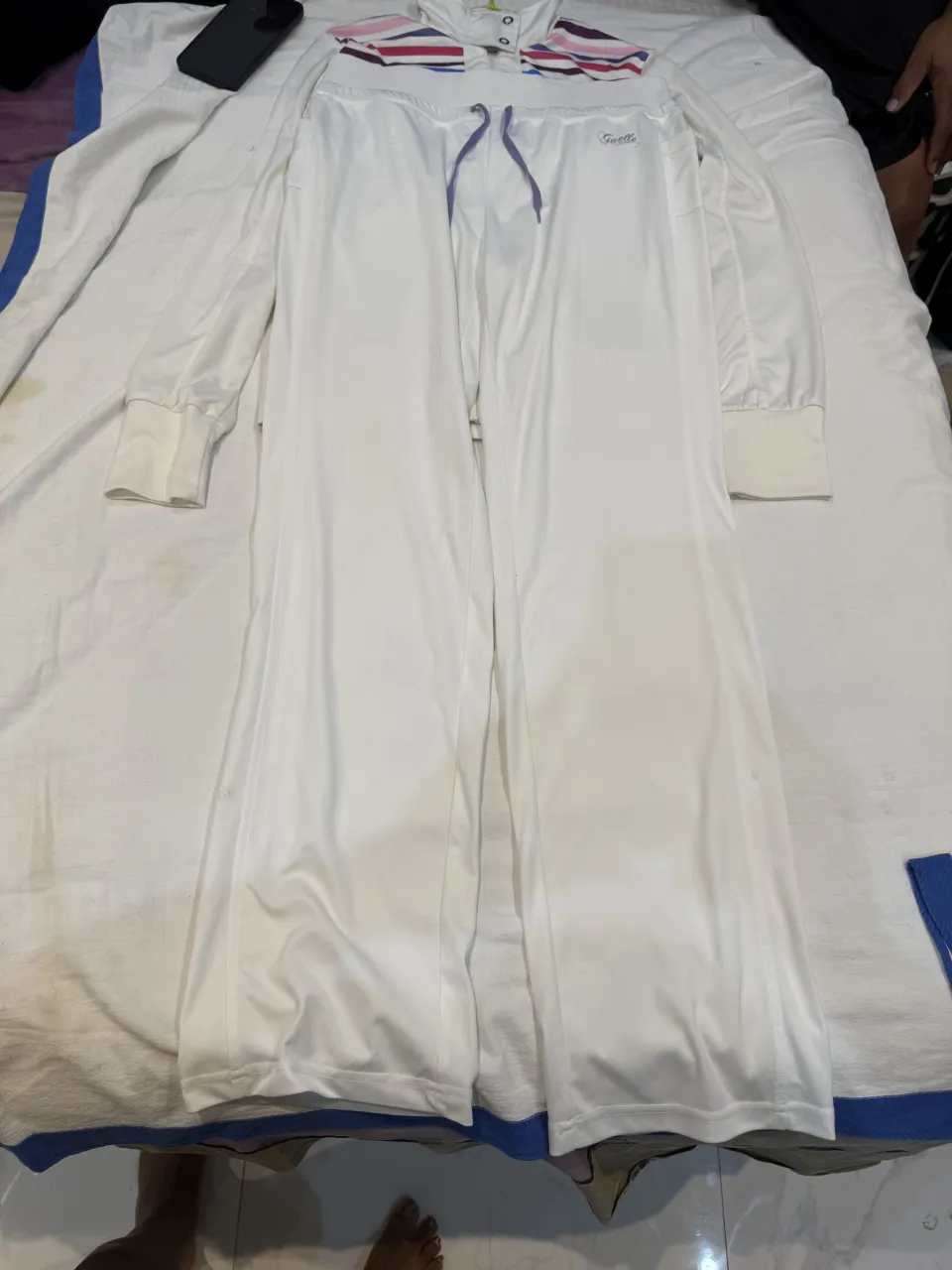 Conjunto deportivo de campera y pantalón blanco. La campera tiene cierre al frente, cuello redondo y detalles de rayas de colores en los hombros. El pantalón es liso con cordón ajustable en la cintura.