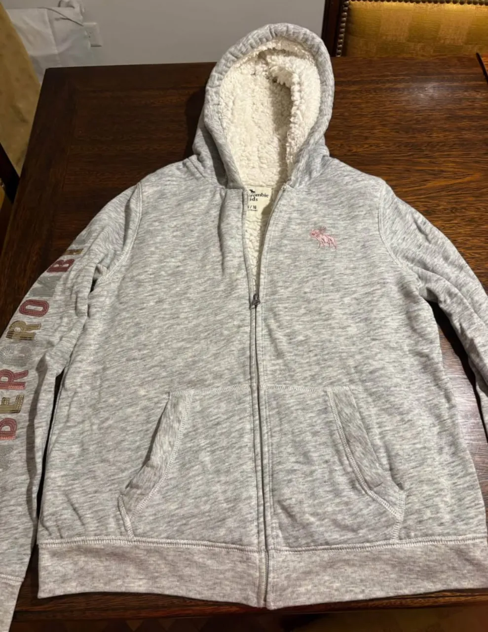 Campera ABERCROMBIE con cierre para mujer, de color gris jaspeado, con capucha forrada en sherpa blanco. Tiene el logo de la marca bordado en rosa en el pecho y detalles en la manga. Ideal para un look casual y abrigado. Excelente estado, muy poco uso. Talle 15-16 KIDS.