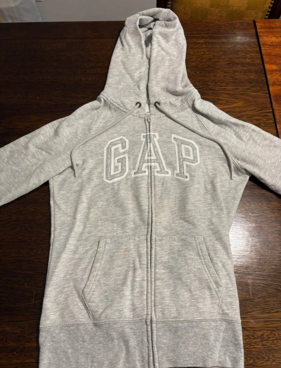 Buzo con cierre y capucha para mujer, de color gris, con el logo de GAP en el frente. Ideal para un look casual y cómodo.