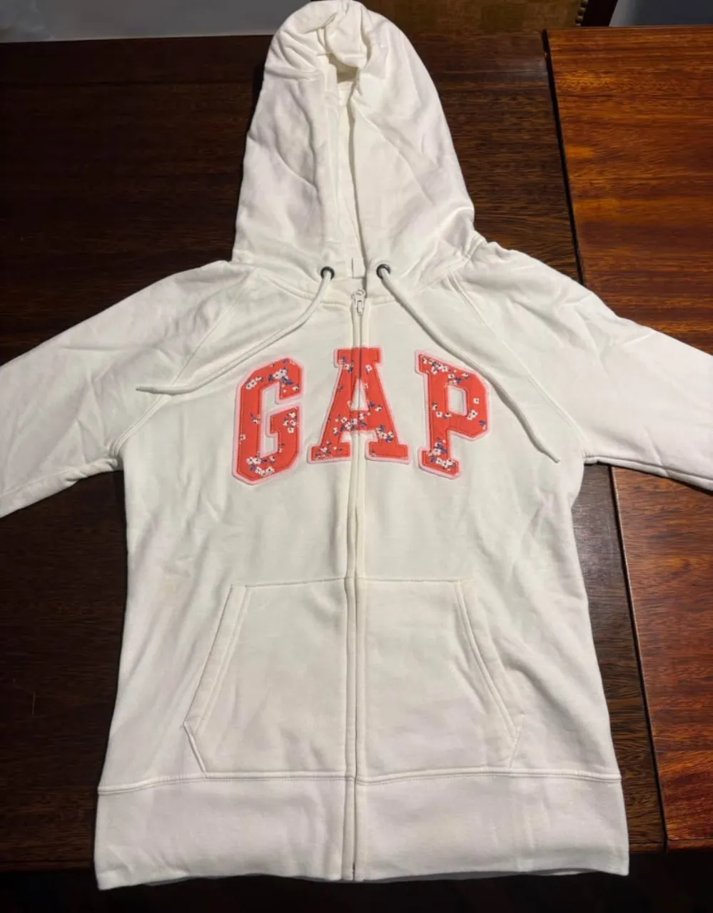Buzo con cierre de la marca GAP, color blanco con el logo GAP en rojo con flores. Ideal para un look casual y cómodo.