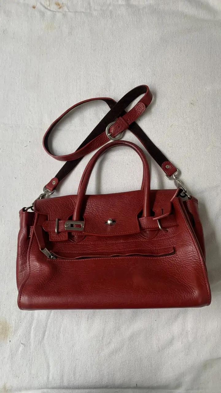 Cartera de cuero roja, con manijas y correa para colgar. Tiene detalles de hebillas y cierre. Ideal para llevar tus pertenencias con estilo.