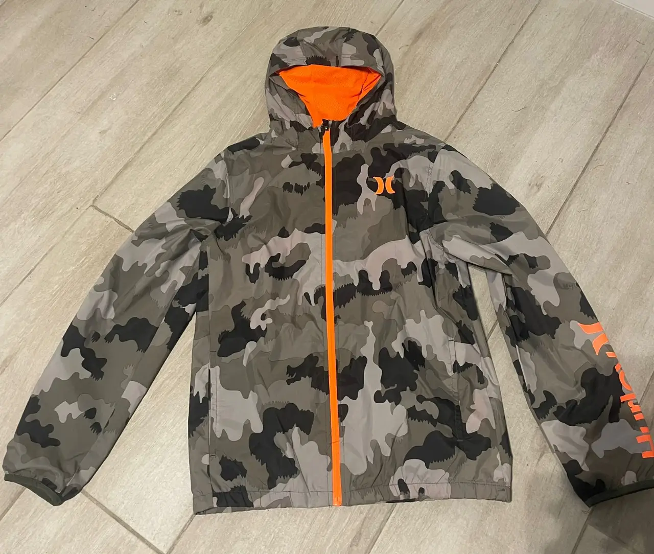 Campera deportiva Hurley con estampado camuflado en tonos grises y negro, con detalles en naranja. Ideal para actividades al aire libre.