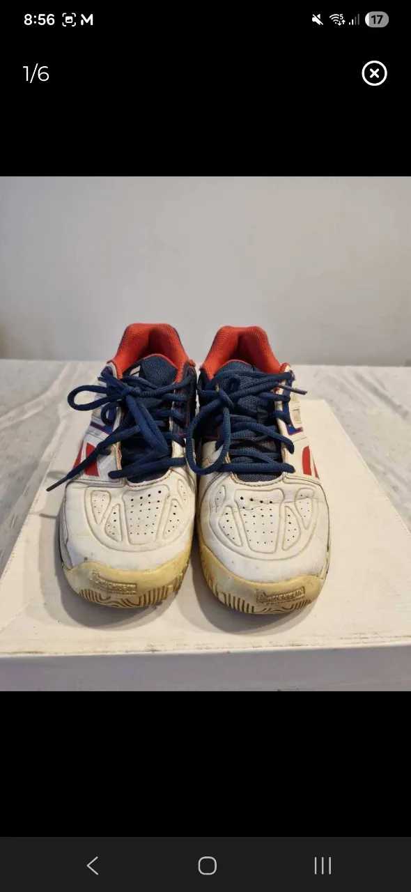 Zapatillas deportivas blancas con detalles en rojo y cordones azules. Presentan un diseño con perforaciones en la punta y un logo lateral.