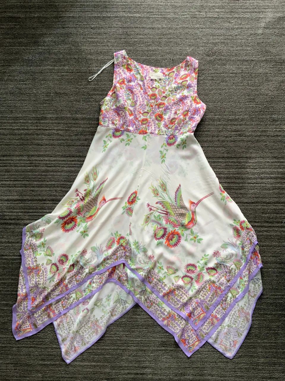 Vestido blanco con estampado floral y de pájaros en tonos vibrantes. Diseño asimétrico con volados en el borde inferior. Escote en V y sin mangas.