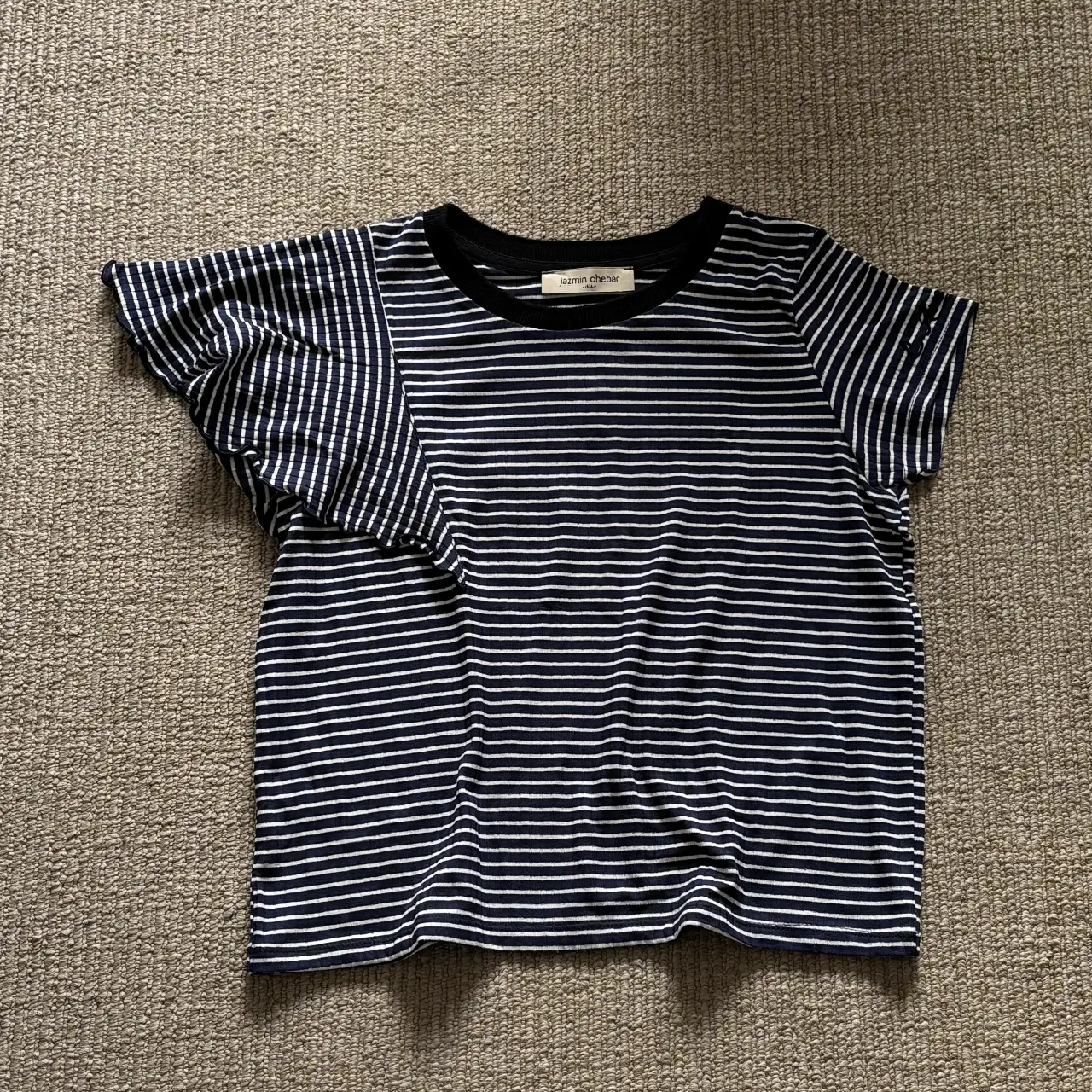 Remera a rayas horizontales en azul y blanco. Tiene mangas cortas con volados y cuello redondo negro. Ideal para un look casual y fresco.