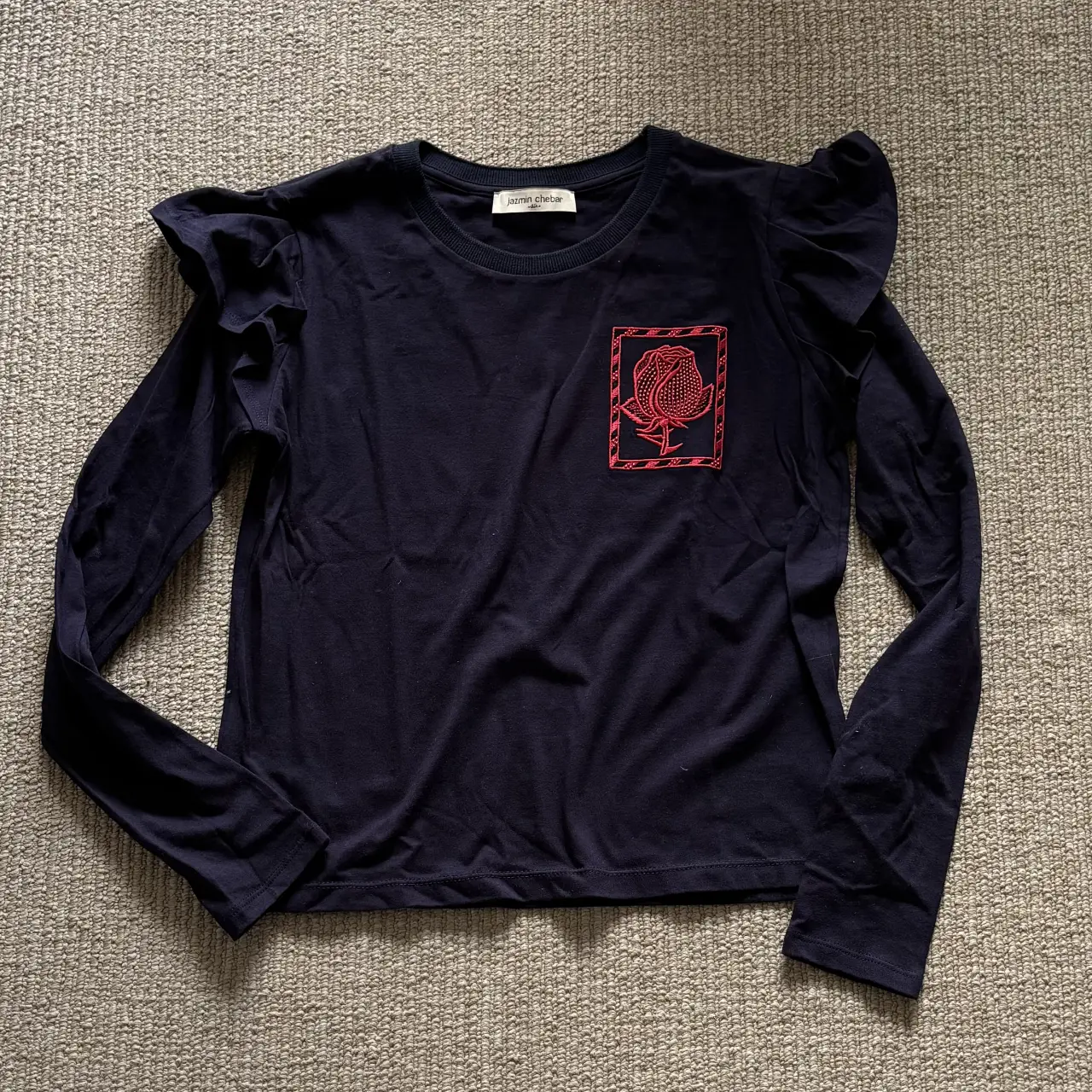 Remera manga larga azul marino con detalle de volados en los hombros y un bordado rojo en el pecho. Ideal para un look casual y moderno.