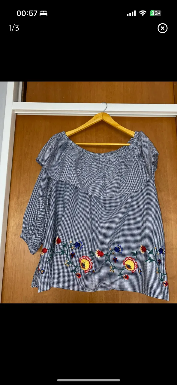 Blusa de mujer con estampado de cuadros vichy en azul y blanco, con detalles florales bordados en la parte inferior. Diseño off-shoulder con volantes en el cuello y mangas 3/4. Ideal para un look casual y fresco.