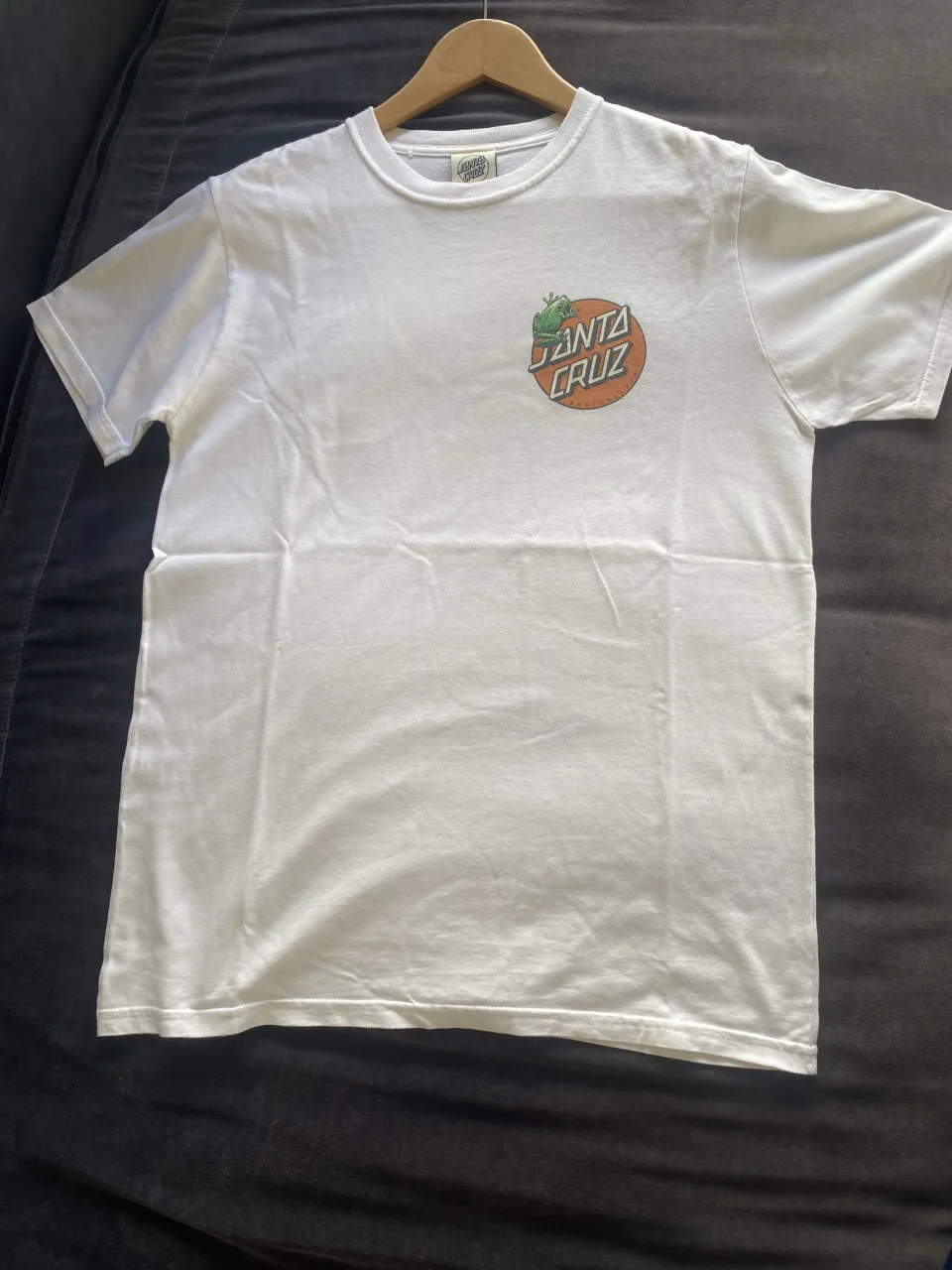 Remera blanca de manga corta con estampa circular en la espalda de "SANTA CRUZ" con flores y hojas. En el frente, sobre el lado izquierdo del pecho, tiene un logo circular de "SANTA CRUZ" con un sol naranja.