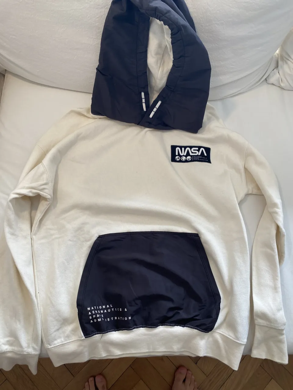 Buzo blanco con capucha negra y bolsillo frontal negro. El bolsillo tiene un logo de la NASA y texto "NATIONAL AERONAUTICS & SPACE ADMINISTRATION".