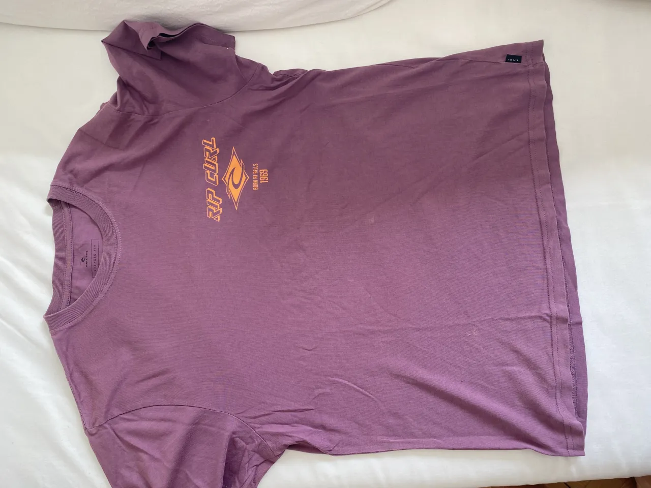 Remera manga corta color lila con estampa "RIP CURL" en naranja en la espalda. Cuello redondo.