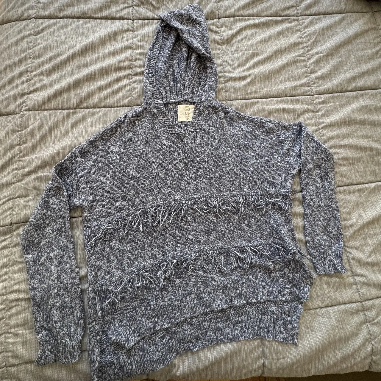 Buzo con capucha de tejido, con flecos en el frente. Es más largo de atrás (53cm de adelante y 60cm de atrás). El material estira. Es de color azul y gris jaspeado, ideal para un look casual y abrigado.