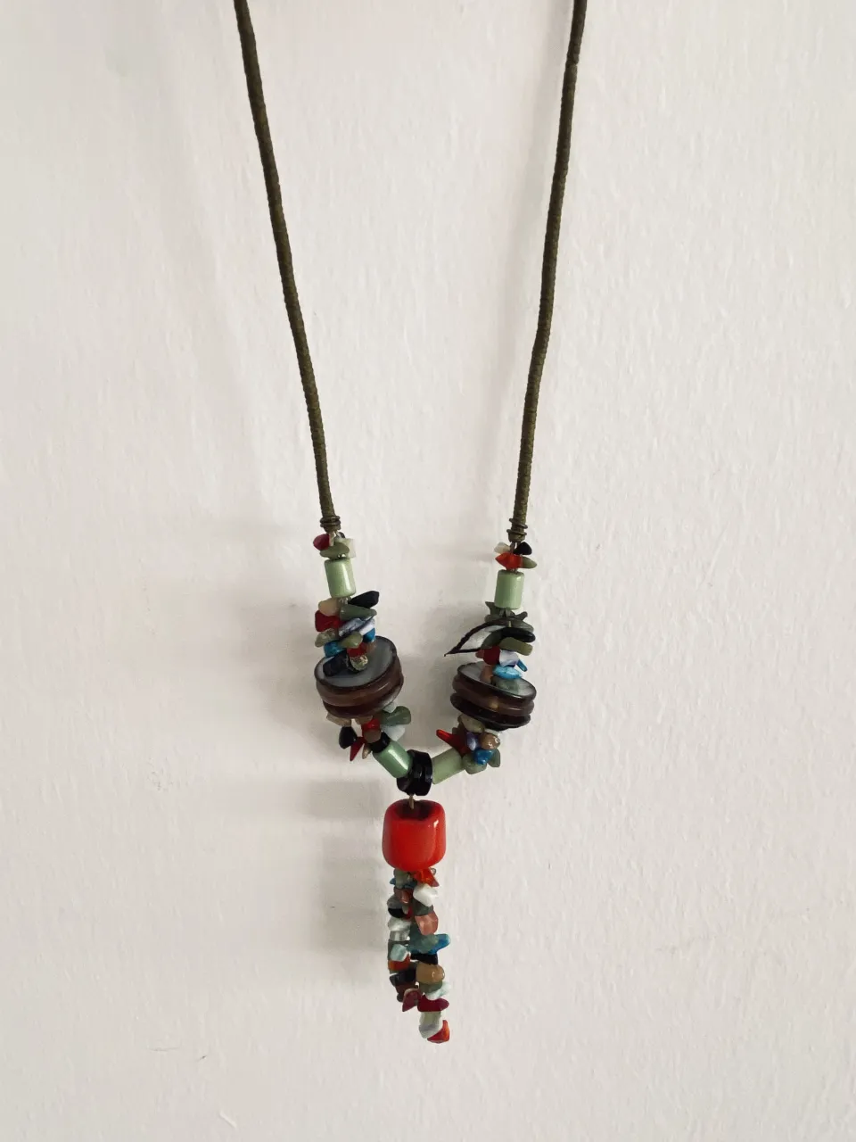 Collar largo con cordón verde y múltiples cuentas de colores, madera y un dije central rojo.