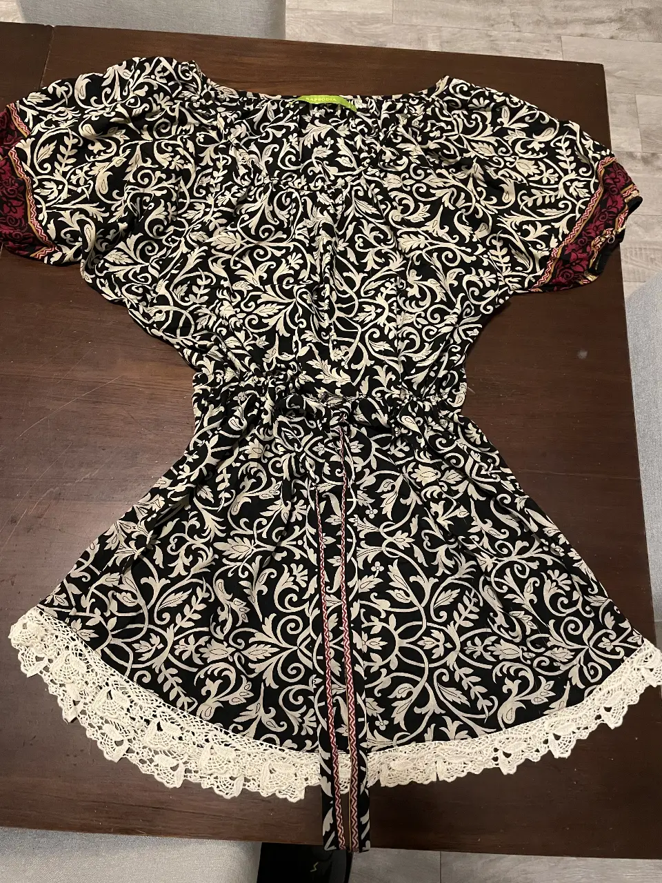 Blusa de mujer con estampado floral en blanco y negro, estilo boho. Tiene mangas cortas con detalles en rojo y blanco, cintura ajustada con cordón y detalles de encaje en el borde inferior. Ideal para un look casual y fresco.