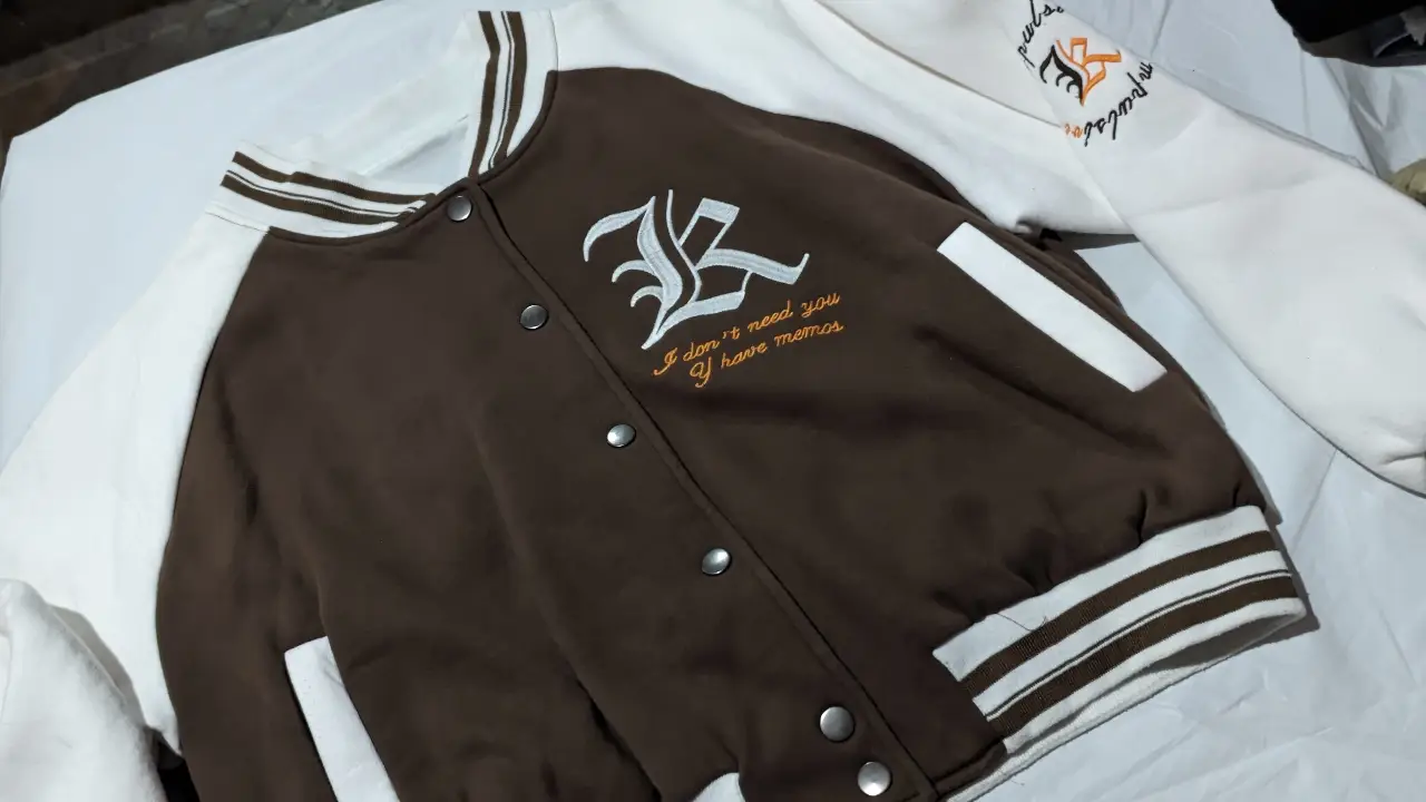 Campera estilo bomber en color marrón con mangas blancas. Posee un bordado en el frente con un diseño y frase. Cierre frontal con botones.