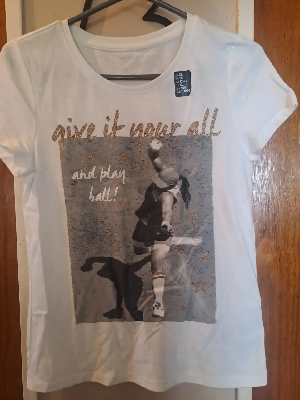 Remera deportiva blanca con estampado de una jugadora de béisbol y la frase 'give it your all and play ball!' en dorado. Ideal para actividades deportivas o uso casual.