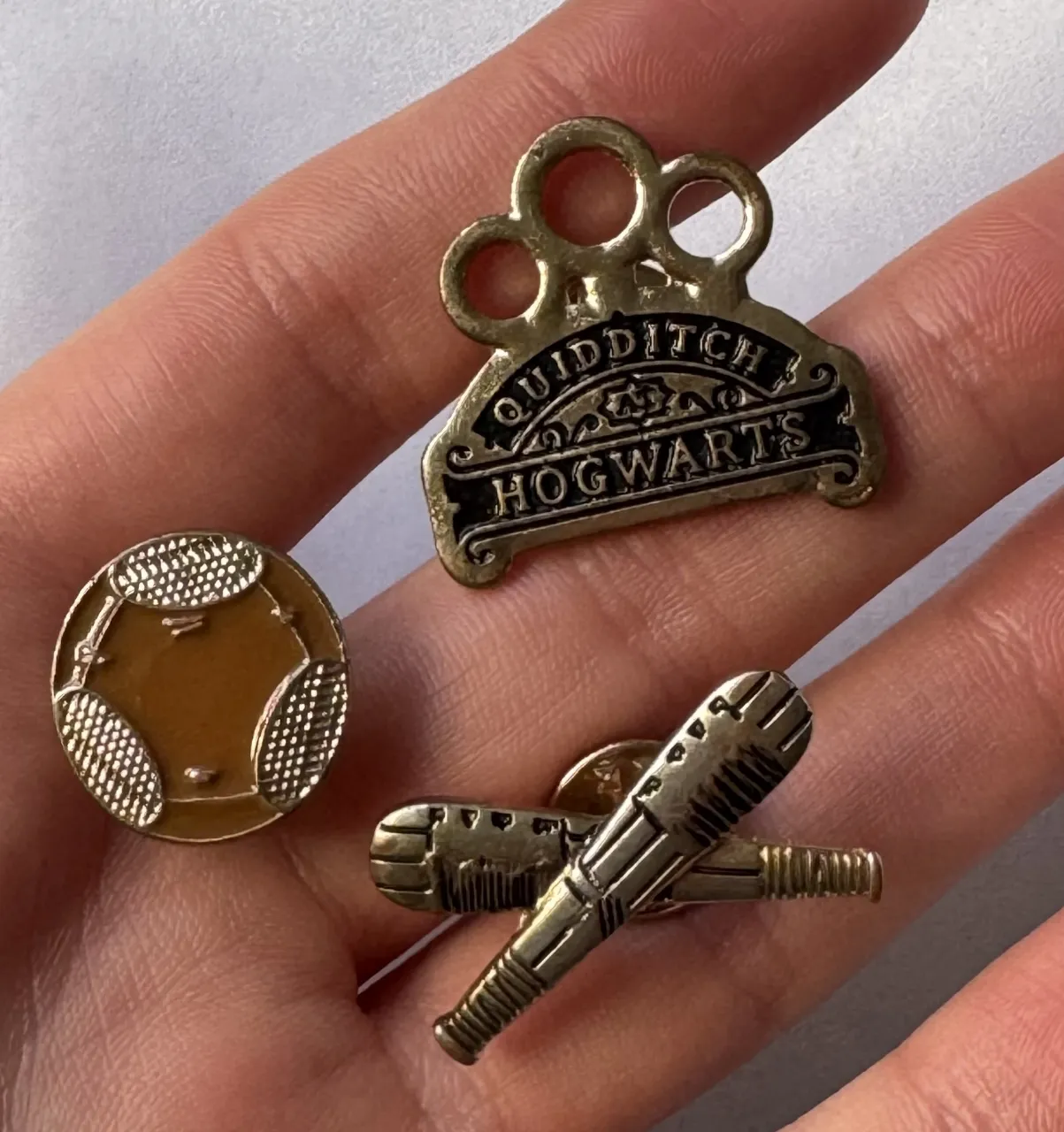Set de 3 pines de metal dorado con motivos de Quidditch de Hogwarts. Incluye un pin con el escudo de Quidditch, otro con dos palos de escoba cruzados y un tercero con una snitch.