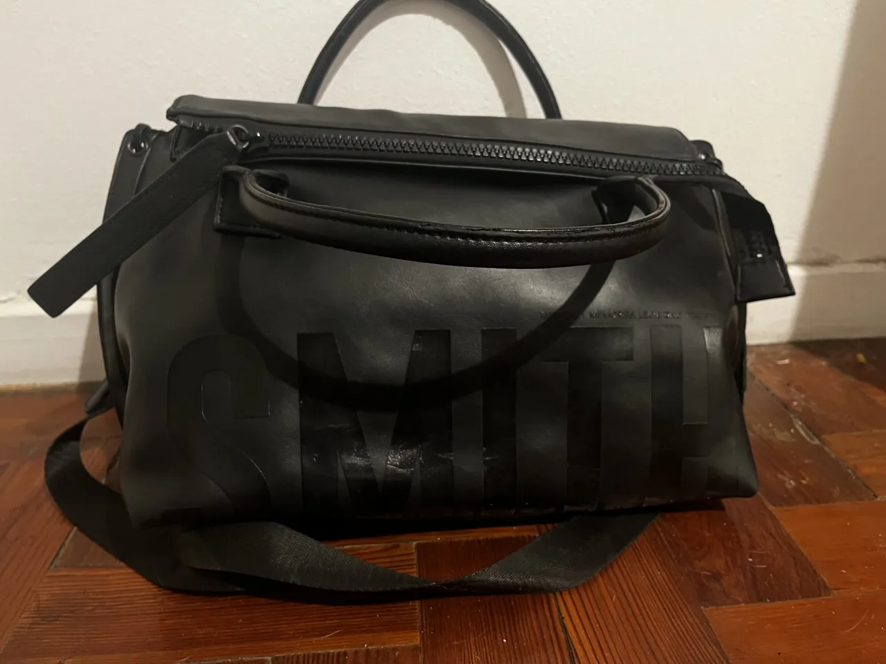 Bolso negro con logo grande en relieve y correa desmontable. Tiene manijas cortas y cierre superior.