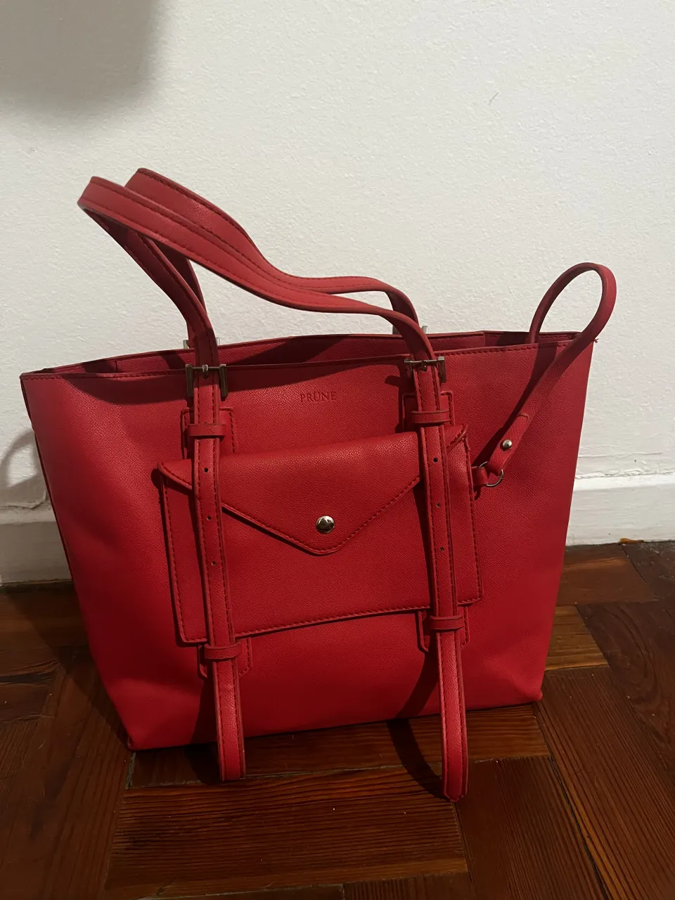 Bolso tote rojo de cuero sintético. Posee un bolsillo frontal con tapa y botón metálico. Las asas son largas y cómodas. La marca "PRUNE" se encuentra grabada en el frente.