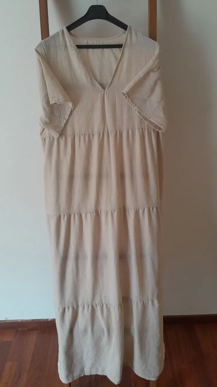 Vestido largo de lino color crudo, con escote en V y mangas cortas. Diseño de tres capas con frunces sutiles, ideal para un look relajado y fresco. Perfecto para días de verano o para estar cómoda en casa.