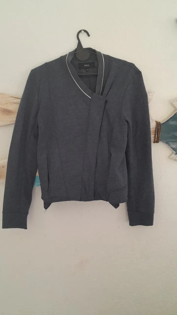 Chaqueta gris con cuello escote en V y detalles de ribetes blancos en el cuello y puños. Posee un cruce asimétrico en el frente.