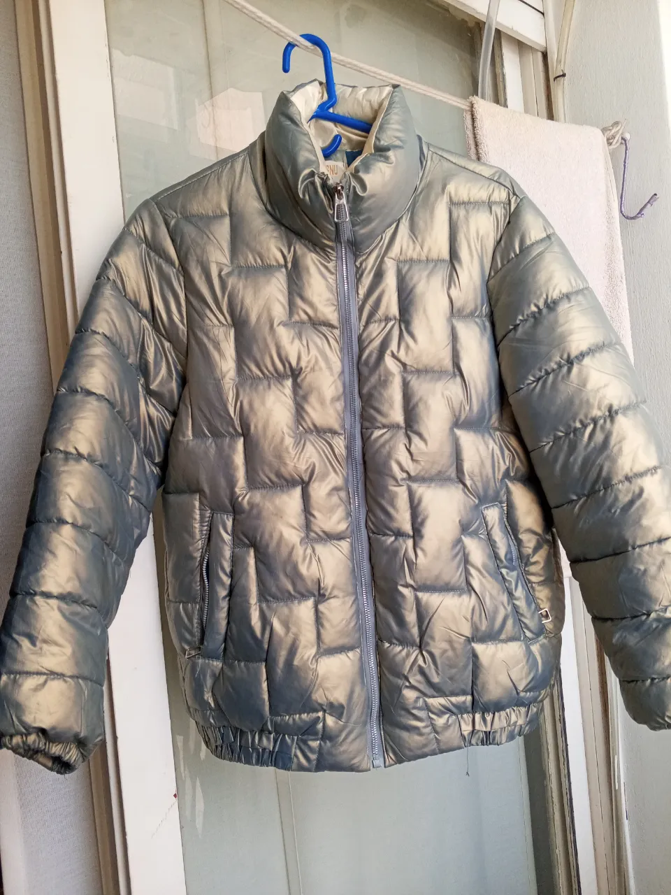 Campera acolchada metalizada con cuello alto y cierre al frente. Posee bolsillos laterales con cierre y elástico en puños y cintura. En la espalda tiene un diseño de estrella formado por las costuras.