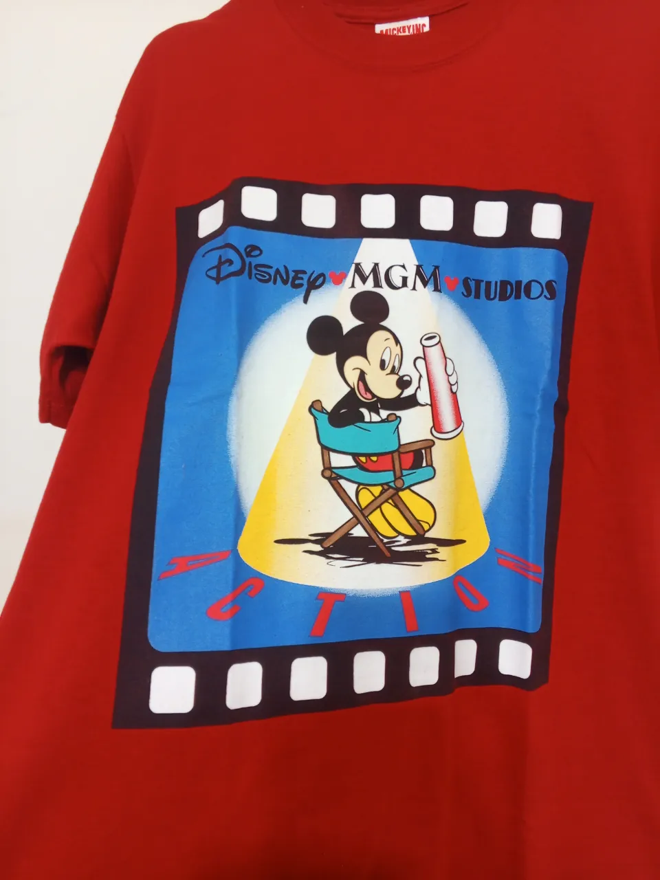 Remera roja de manga corta con estampa de Mickey Mouse en el frente. La estampa muestra a Mickey sentado en una silla de director, con el logo de Disney MGM Studios. Ideal para fans de Disney y para un look casual y divertido.